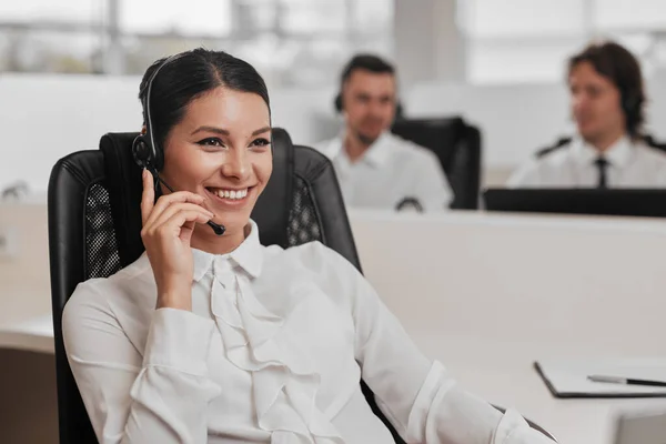 Friendly Hispanic woman in white blouse adjusting headset microphone and smiling, while sitting on chair and speaking with client during work in call center office