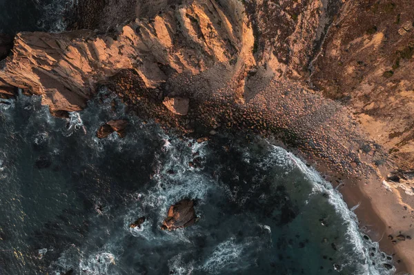 Top view from drone of foamy waves running on rocky shoreline with cliffs in sunlight