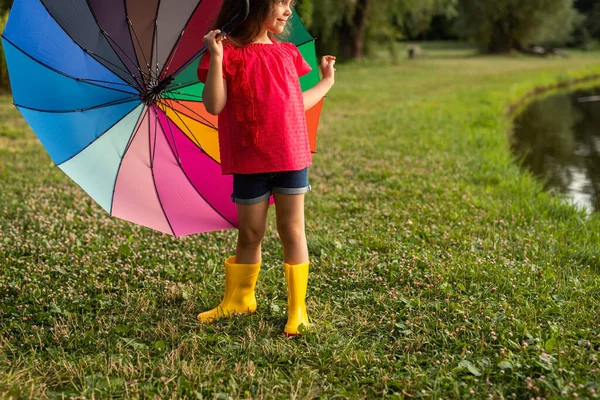 身穿时髦服装 头戴黄色胶靴的黑头发的积极的小女孩 站在池塘边的草地上 手里拿着五彩缤纷的雨伞 面带微笑地望着远方 — 图库照片