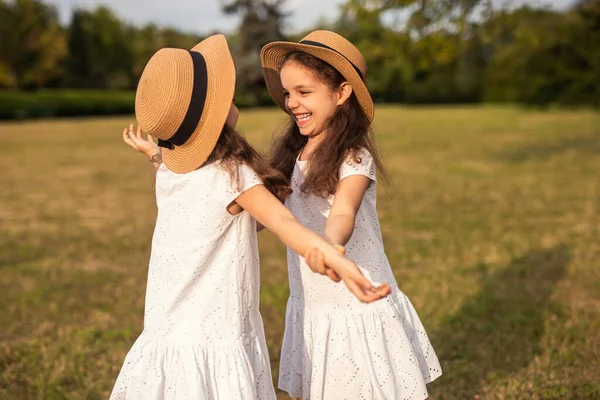 同じような白いドレスとわらの帽子で長い暗い髪を持つ愛らしい陽気な小さな双子の女の子 田舎の休日の間に草の牧草地で一緒に遊んでいる間に明るく笑顔 — ストック写真