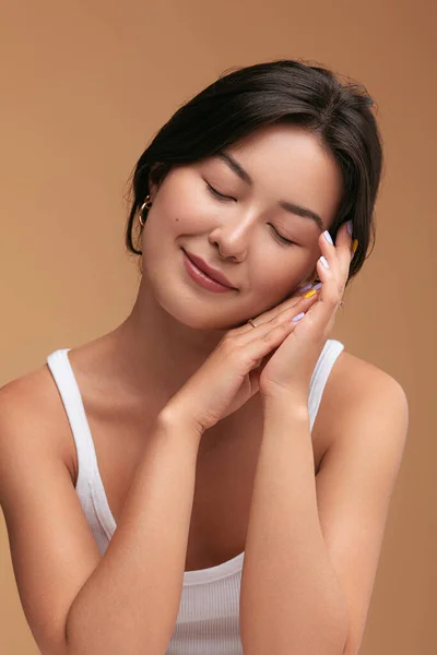 在棕色背景下的日常美容过程中 年轻的亚洲女性合上眼睛 触摸柔软的面部和手部肌肤 令她们感到高兴 — 图库照片