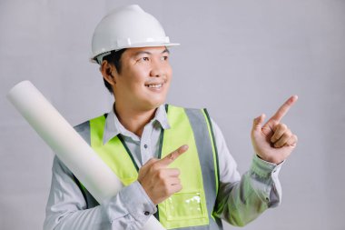 Bir mimar ya da mühendis, reflektör ve inşaat işi için kaskı olan genç bir adam, el kol hareketi yaparken ve parmağını uzaya doğrulturken bir tasarım planı tutuyor..