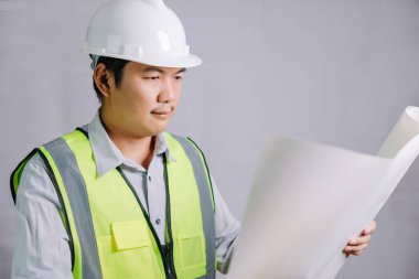 Yansıtıcı takım elbise giyen genç bir mimar ya da mühendis ve inşaat için güvenlik kaskı gri bir arkaplanda tasarımının taslağını tutuyor..