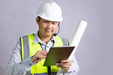 Bir mimar ya da mühendis, yansıtıcı takım elbise giyen ve inşaat için kaskı olan genç bir adam, tablet üzerinde eline dokunurken bir tasarım projesinin planını tutuyor..