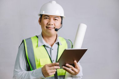 İnşaat işleri için reflektör ve kaskı olan genç bir adam, mimar ya da mühendis elinde tabletle elinde tasarım planıyla kameraya bakıyor..