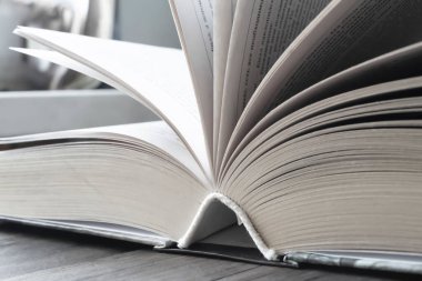Açık kitap. Kitabın yaprakları farklı yönlere uzanıyor. Sessiz renklerle çekilmiş bir fotoğraf. Masanın üzerindeki kalın kitap açık..