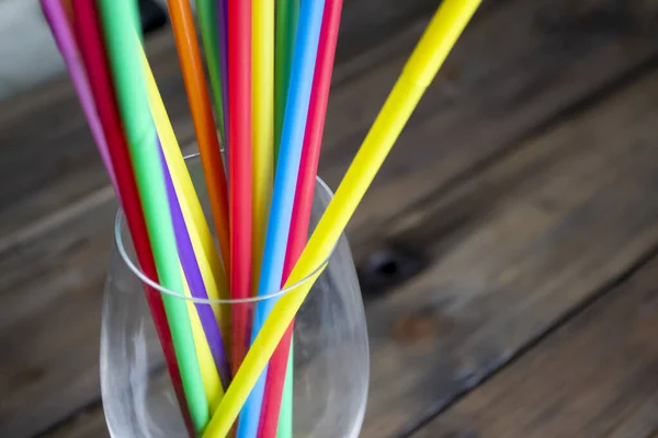 Plastic cocktail straws in a glass glass on a wooden table. The background is a tree, there are many colored straws for drinks in the glass.