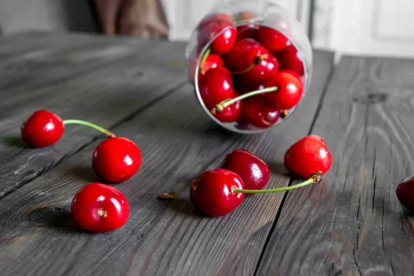 ON the wooden table is a glass of cherry berries. Cherry berries from a lying glass are scattered on the table. Wooden table. A glass of cherry berries.