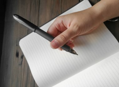 There is a black pen in the mans hand. The man holds a black pen over an open notebook. A blank notebook for notes and a mans hand with a pen.