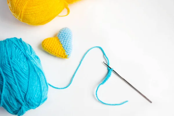 A knitted heart with a crouch. Crocheting the heart. On a white background, a hook, threads and a two-tone heart are the final work. The heart of the threads is the flag of Ukraine.