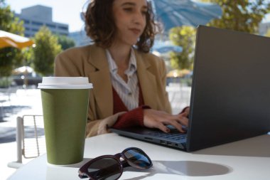 Seçici odak noktası kahve ve güneş gözlükleri, arka planda dizüstü bilgisayarda çalışan Latin kadın, dijital göçebe konsepti.
