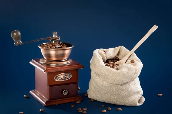 Grãos de café em um moedor de café manual em um fundo azul escuro. Café num saco de lona. Conceito. — Fotografia de Stock