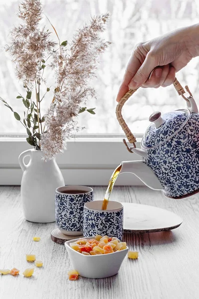 Bir el çaydanlıktan çay doldurur. Çay zamanı. Beyaz seramik vazoda kuru tahıl dalları, küçük mavi desenli çay seti, şekerli meyveler.. 