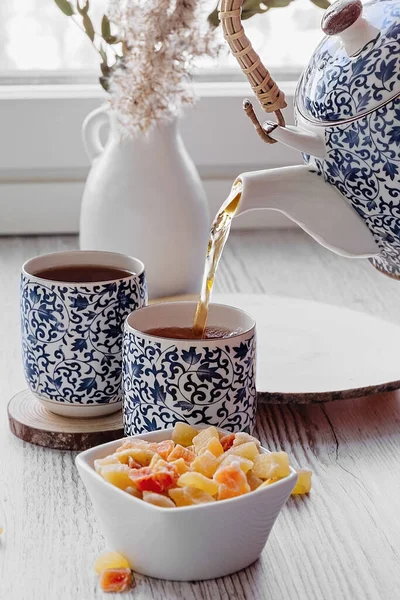 Sebuah tangan menuangkan teh dari teko teh. Waktunya minum teh. Cabang sereal kering di vas keramik putih, satu set teh dengan pola biru kecil, buah manis. — Stok Foto