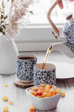 Bir el çaydanlıktan çay doldurur. Çay zamanı. Beyaz seramik vazoda kuru tahıl dalları, küçük mavi desenli çay seti, şekerli meyveler.. 