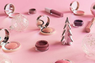 Güzellik ürünleri, Noel baloları ve pembe arka planda dekoratif Noel ağacı. Kızarmalar, göz farı ve dudak parlatıcısı. Noel partisi makyaj konsepti