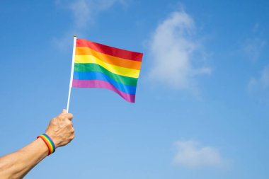 Adamın eli LGBT gökkuşağı bilekliği takıyor. Arka planda LGBT gökkuşağı bayrağı sallıyor. LGBTQ + eşitlik hareketi topluluğu kavramı.