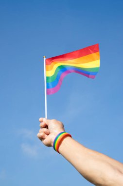 Adamın eli LGBT gökkuşağı bilekliği takıyor ve arka plan mavi gökyüzünde LGBT gökkuşağı bayrağı sallıyor (seçici odak), LGBTQ + eşitlik hareketi topluluğu kavramı