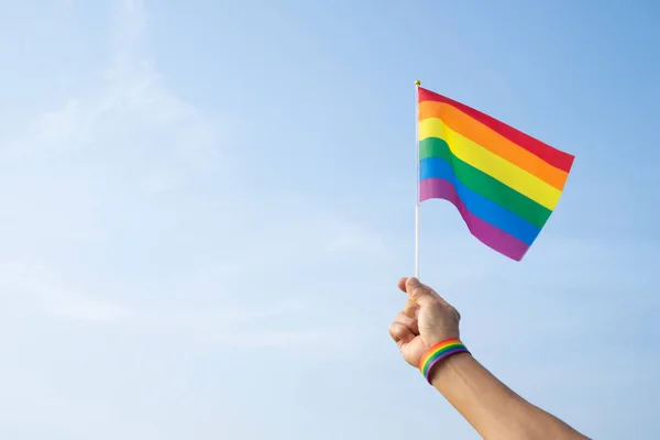 Adamın eli LGBT gökkuşağı bilekliği takıyor ve arka plan mavi gökyüzünde LGBT gökkuşağı bayrağı sallıyor (seçici odak), LGBTQ + eşitlik hareketi topluluğu kavramı