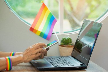 Üst düzey kadın elleri, gökkuşağı bilekliği takıyor. Akıllı telefon, Igbt bayrağı ve dizüstü bilgisayar kullanıyor. LGBTQ eşitlik hareketini destekleyen teknoloji kavramı, LGBTQ mutlu gurur ayı