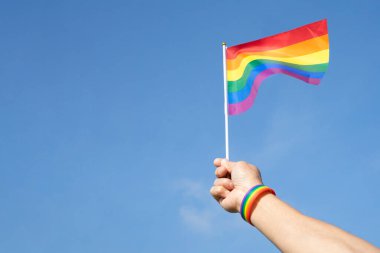 Adamın eli gökkuşağı bilekliği takıyor. Mavi gökyüzünde LGBT gökkuşağı bayrağı sallıyor. Kopya alanı, LGBTQ + toplum eşitliği hareketi konsepti, LGBTQ + mutlu gurur ayı.
