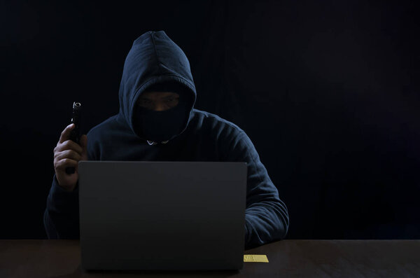 Хакер в воровской толстовке с пистолетом взламывает компьютерную систему на ноутбуке, концепцию киберпреступлений