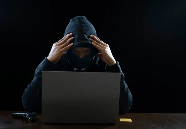 a stress hacker in black hoodie doing both hands on head is hacking failed,there are device such as gun,credit card and laptop on table isolated on dark background