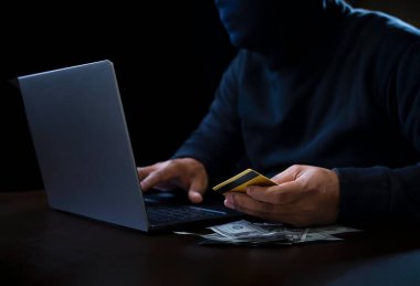 concept of internet theft ,cyber crime., hacker in black hoodie and thief mask using laptop to hack credit card, a lot of money on desk