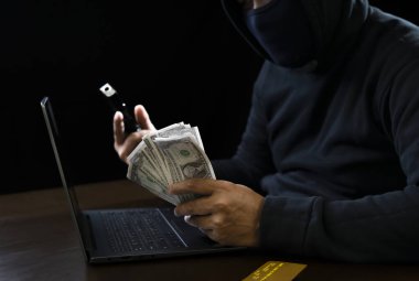 Siber suçlar konsepti, siyah kapüşonlu soyguncu elinde para ve silah, kredi kartıyla karanlık arka planda duruyor.