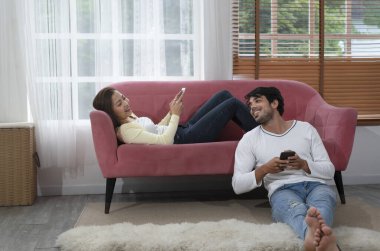 Genç çift oturma odasında dinlenirken akıllı telefon oynuyor. Aile hayatı teknolojisi, hafta sonları eğlence.