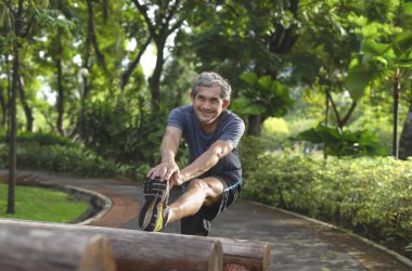 Yaşlı bir adam koşmadan önce parkta bacaklarını açık havada spor malzemelerine uzatıyor. Yaşlı insanlar ısınıyor, sakatlığı önlemek için egzersiz yapmadan önce vücudunu esnetiyor.