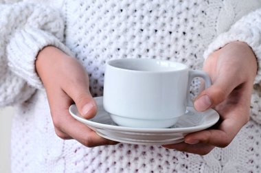 Beyaz fincan ve çay tabağı tutan, sıcak örülmüş beyaz kazaklı bir kızın ellerini yakın plan çek. Kış konforu, rahatlama kavramı