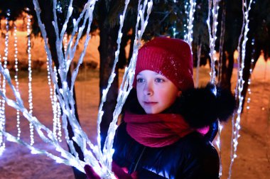 Kırmızı şapkalı ve eşarplı güzel bir kız Noel ışıkları çelenklerinin arkasında duruyor. Yeni yılın şenlik havası
