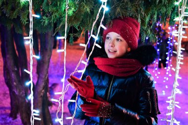 Kırmızı şapkalı ve eşarplı güzel bir kız Noel ağacı ve ışık çelenklerinin arkasında duruyor. Yeni yılın şenlik havası