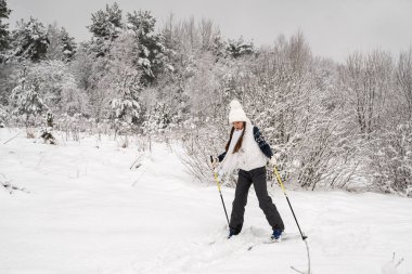 Beyaz şapkalı ve yelekli bir kız kışın karla kaplı ağaçların arasında kayak yapmaya gider. Aile aktiviteleri, kış sporları.
