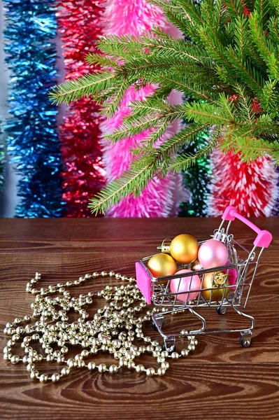 クリスマスの装飾と光沢のあるボールでいっぱいの装飾的な食料品のカートは 多色のチンゼルと緑のトウヒの枝を背景に立っています 新年とクリスマスのショッピングの概念 — ストック写真