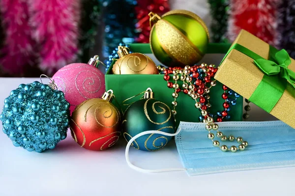 医療マスクは美しいクリスマスの装飾の隣にあります 多色のティンセルを背景に箱の中のおもちゃ ビーズ ボール パンデミックと隔離における新年 — ストック写真