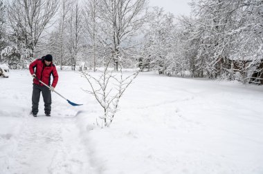 Kırmızı ceketli adam köyde kürekle kardan yolu temizliyor. Kış endişeleri