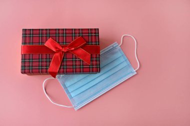 Tıbbi koruma maskesi ve pembe arka planda kırmızı hediye kutusu. Üst Manzara. Virüslerin bayram kutlamalarındaki etkisi. Boşluğu kopyala