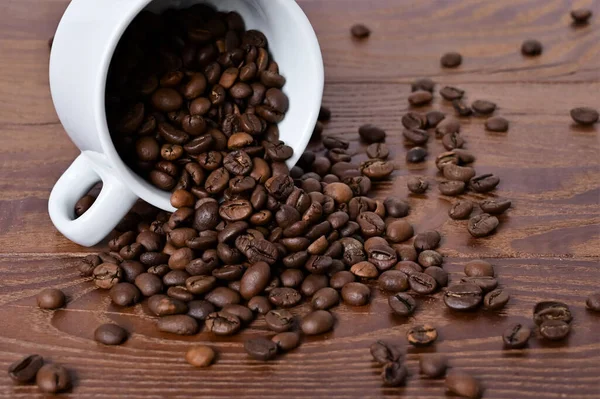 コーヒー豆は白いカップから木製のテーブルにこぼれる 木の背景にコーヒー豆 — ストック写真