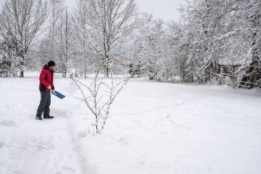 Kırmızı ceketli bir adam köyde karlı yolu kürekle temizliyor.