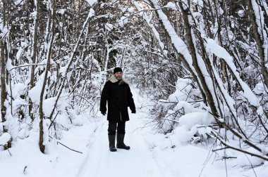 brutal man with beard in warm clothes is standing in winter forest. Person enjoys a walk in fresh air. Peace and quiet in snowy forest