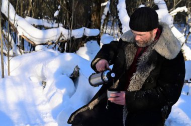 Portrait of brutal man with beard. Adult man in warm sheepskin coat pours hot tea from thermos into cup. Outdoor recreation on frosty winter sunny day in forest