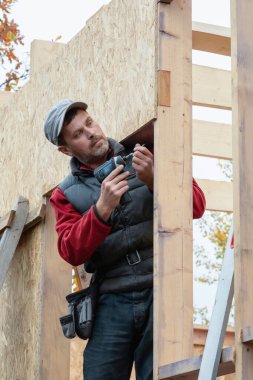 Elinde elektrikli tornavidayla inşaatçının portresi. Erkek işçi tornavidayla ahşap yapı inşa eder. Müteahhit evin ahşap çerçevesini monte ediyor. Sanayi teması