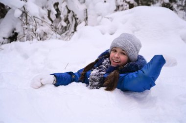 Neşeli güzel kız kar yığınında oturuyor, gülüyor ve elini sallıyor. Kış ormanlarında açık hava aktiviteleri