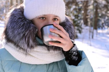 Sıcak giysili güzel kız kış ormanlarında termostan çay içer. Soğuk ve güneşli bir günde açık hava eğlencesi. Sıcak içecek seni ısıtır. Yakın plan.