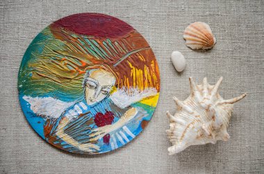 Ellerinde kalbi olan bir melek yuvarlak bir tahtaya akrilikle resmedilmiş. Keten bir çekiçle deniz kabuğunun hayatı.