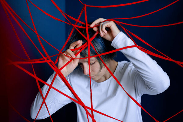 Young woman is entangled in red threads on blue walls background. Concept of obsessive states, paranoia, mental illness.