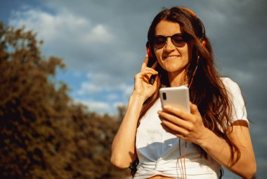 Güzel, mutlu, genç bir kadın cep telefonu, kulaklık kullanıyor ve şehir sokaklarında müzik dinliyor. Pozitif duygular, yaz aylarında hayat tarzı anlamlı portreler..
