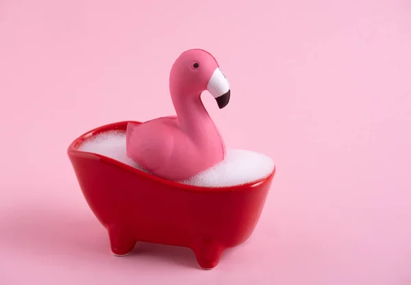 Funny pink flamingo in red bath with foam on a pink background.